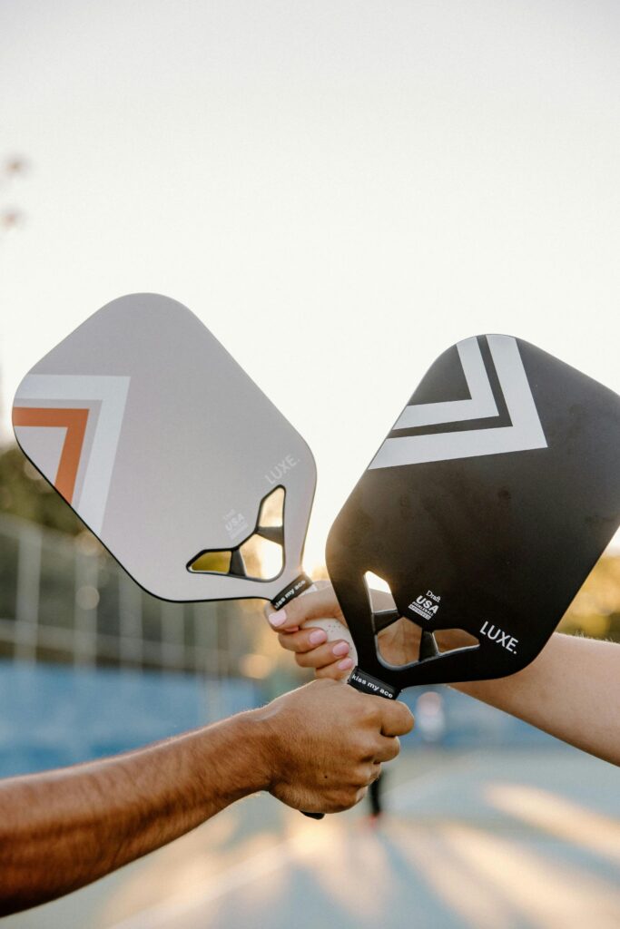 pexels-photo-29775349-29775349 Two hands holding pickleball paddles outdoors during sunset.